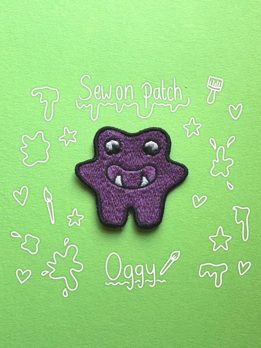 A purple monster-shaped embroidered patch with white eyes and fangs, placed on a bright green background with white doodle-style text reading 'Sew on Patch' and 'Oggy'.