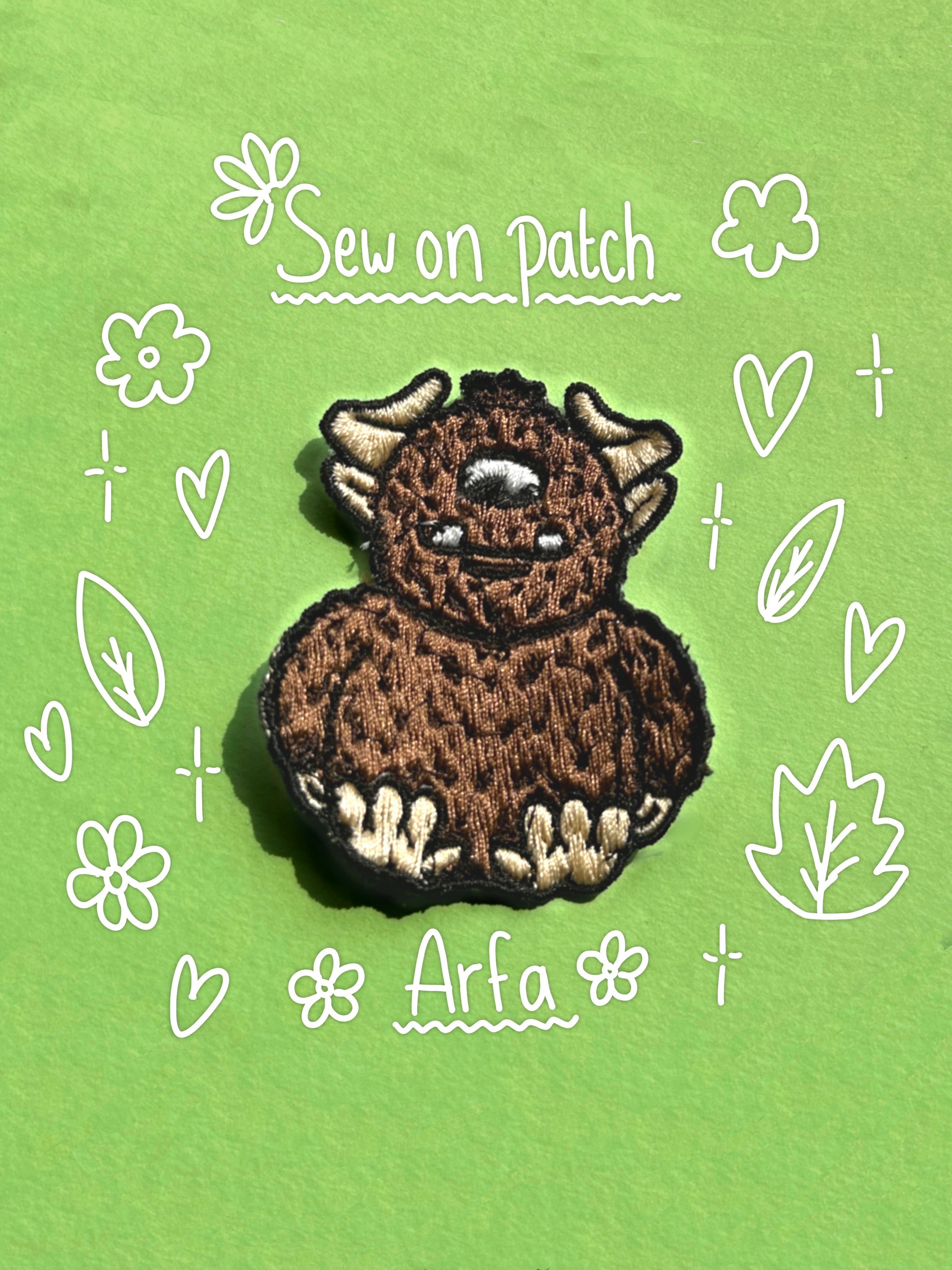 A brown embroidered patch of a cute furry monster with horns and white paws, placed on a green background with white doodle-style text reading 'Sew on Patch' and 'Arfa'.
