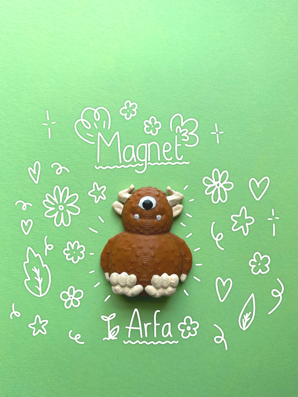 A handmade brown and white  3D magnet shaped like a cute monster, displayed on a light green background with white hand-drawn doodles and the words 'Magnet' and 'Arfa' with flower and leaf doodles surrounding it.