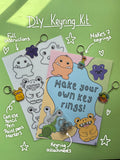 DIY keyring kit image shows frosted shrink plastic sheet with monster images printed on, ready to colour in. With instructions sheet and the keyring attachments that come in the kit. Display keyrings surround the items with white hand written notes and doodles detailing the artist materials that can be used including pencil, pen, paint pens and alcohol markers. 
