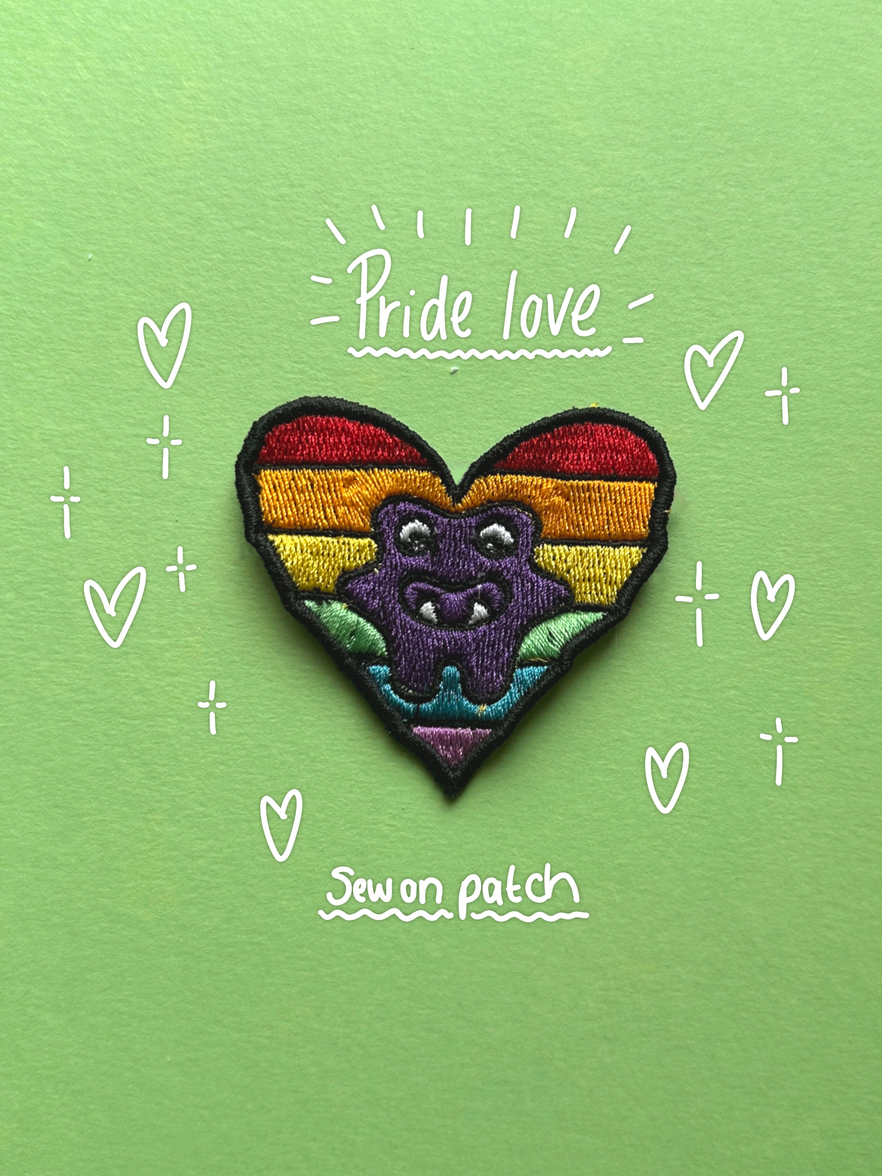 A heart-shaped embroidered patch featuring a purple monster on a rainbow-striped background, displayed on a light green surface with hand-drawn white hearts and the words 'Pride love' and 'Sew on patch'