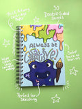 A spiral-bound sketchbook with a colorful cover illustration of a purple monster holding a paintbrush and the words 'Always be creative!' on a light green background with handwritten callouts.