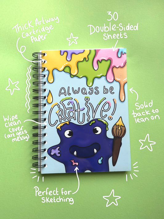 A spiral-bound sketchbook with a colorful cover illustration of a purple monster holding a paintbrush and the words 'Always be creative!' on a light green background with handwritten callouts.