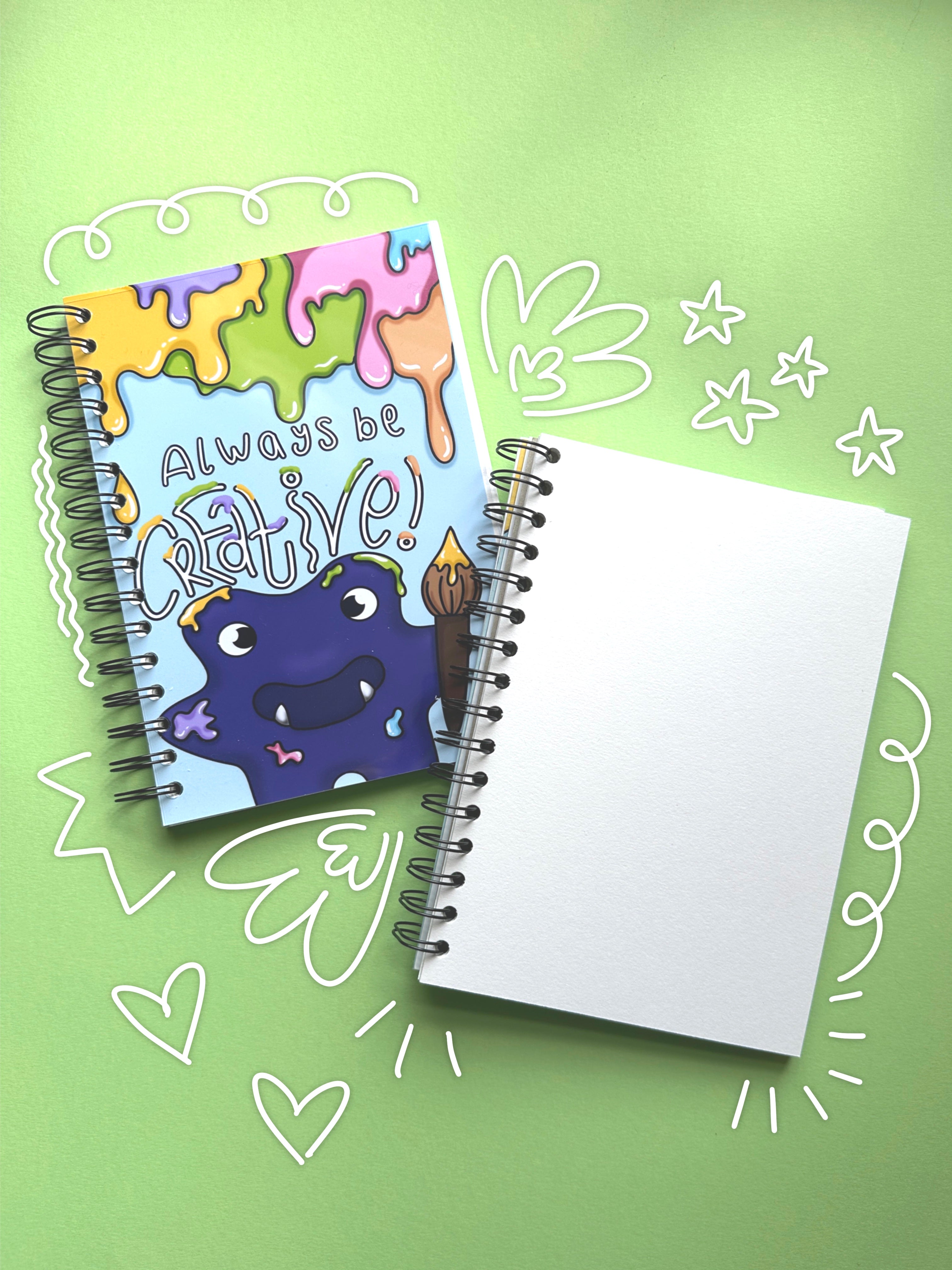 Two spiral-bound sketchbooks on a light green background with white hand-drawn doodle outlines. The left sketchbook has a colourful cover illustration of a purple monster and the text 'Always be creative!'. The right sketchbook is folded back to showcase the blank high quality cartridge paper inside the sketchbook.
