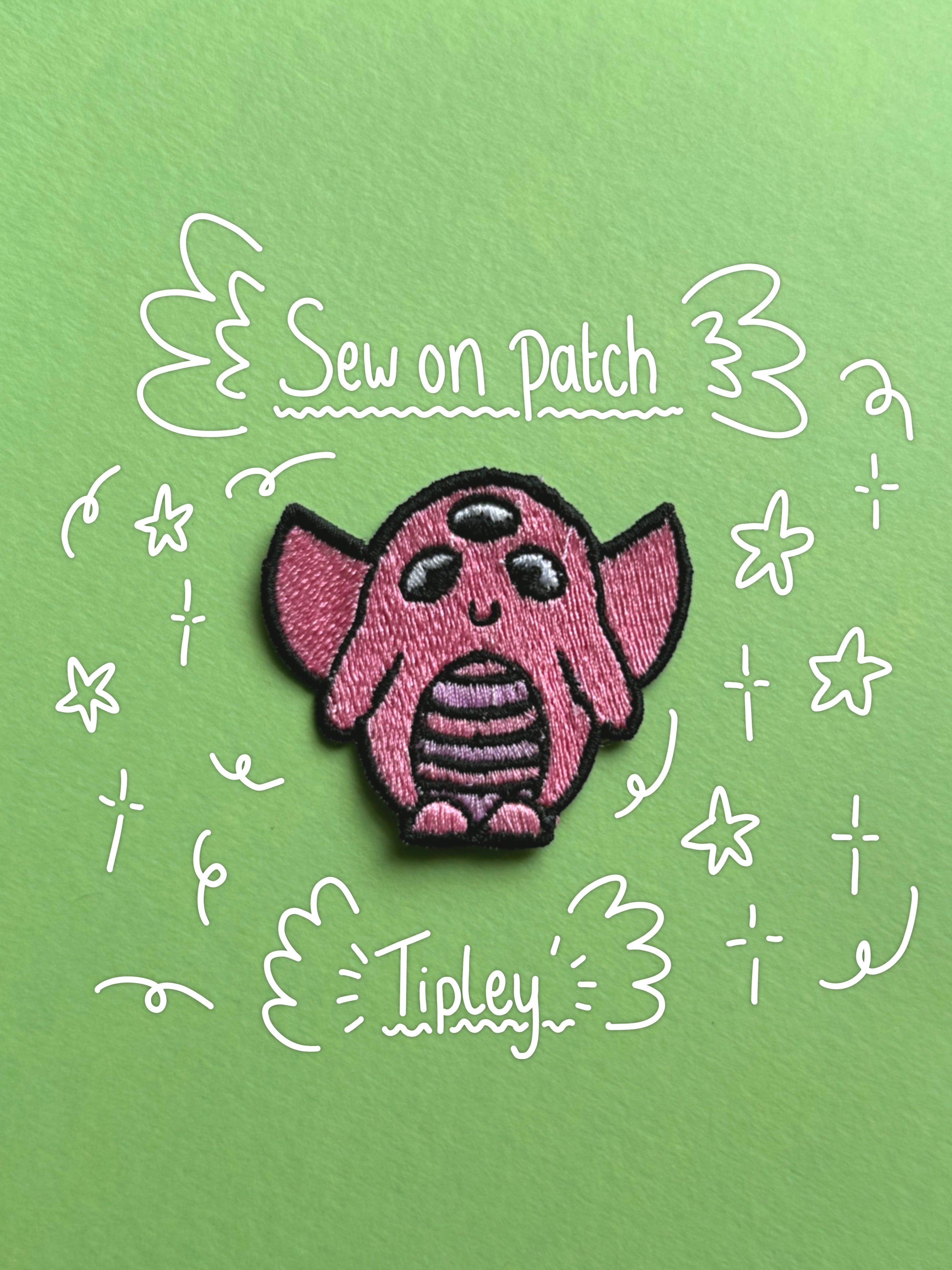 A pink embroidered patch shaped like a three-eyed alien or creature with large ears, centered on a light green background with white hand-drawn doodles and the words 'Sew on Patch' and 'Tipley'.